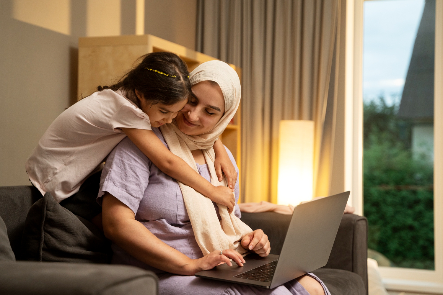 An Arab woman sits on a sofa with a laptop in her arms and is studying something, with a little girl hugging her from behind.